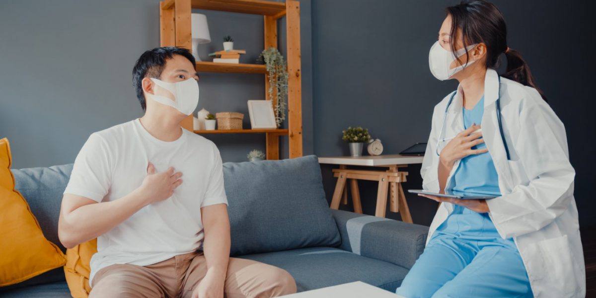 Young Asia female physician doctor wear face mask using digital tablet sharing good health test news with happy male patient sit on couch in house. Medical insurance, Visit patient at home concept.